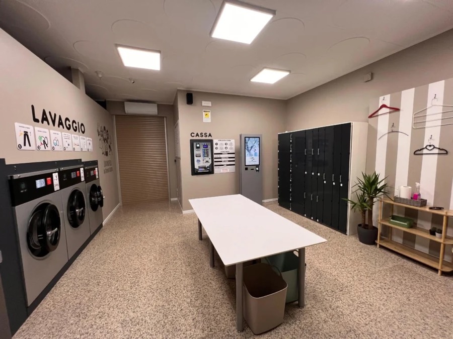 Una lavanderia self service con lavatrici professionali, tavolo centrale e la parete di armadietti Lavasecco Point sul fondo