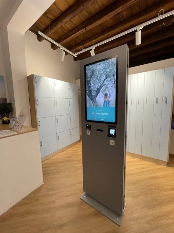 Il totem digitale di un Lavasecco Point, alto un metro e ottanta, con lo schermo verticale acceso e la parete di armadietti alle spalle
